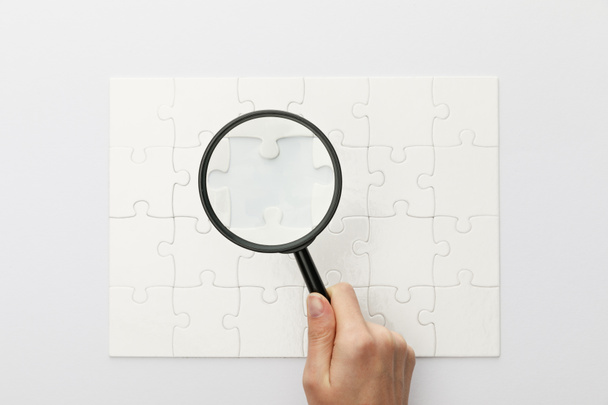 cropped view of woman holding magnifying glass under jigsaw puzzle on white background - Photo, Image