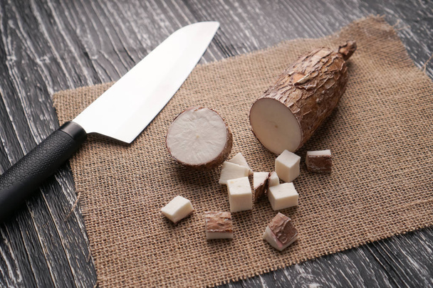 Raíz de yuca en rodajas sobre un saco sobre una mesa de madera negra con un cuchillo
 - Foto, imagen