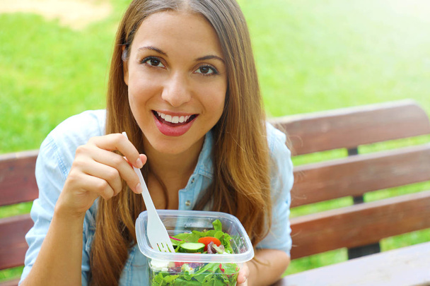 Heureuse femme souriante manger de la salade assis dans le parc regarde la caméra avec espace de copie. Déjeuner salade à emporter conteneur saine alimentation jeune femme d'affaires prête à manger dans le parc mode de vie
. - Photo, image
