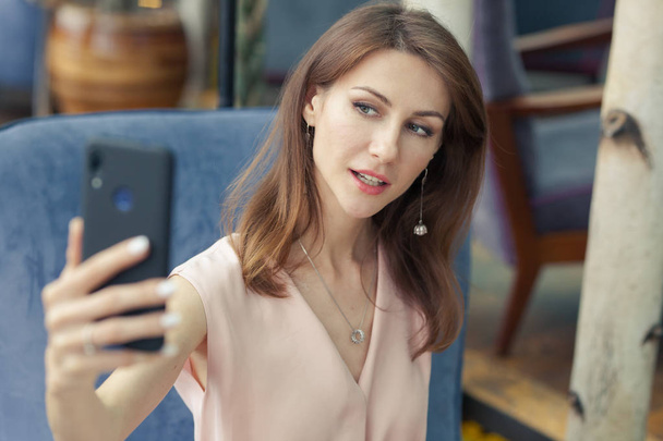 Portrait of attractive smiling young brunette woman in a pink dress sitting in a blue armchair. girl holds a mobile phone in hand and takes a selfie. - Fotó, kép