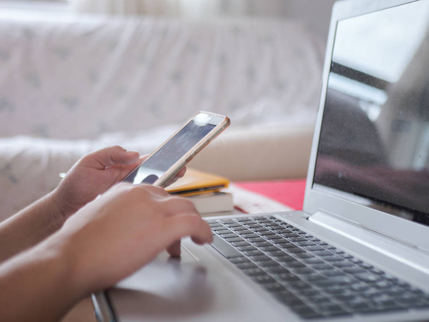 Νεαρή γυναίκα συνεργάζεται με το λευκό smartphone και laptop την ίδια στιγμή από το σπίτι - Φωτογραφία, εικόνα