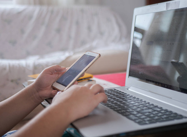 Mujer joven trabaja con su teléfono inteligente blanco y portátil al mismo tiempo desde casa
 - Foto, Imagen
