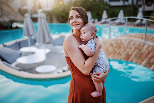 Ritratto di bella bruna caucasica sorridente che tiene il suo amorevole bambino di 6 mesi in piedi accanto alla piscina
. - Foto, immagini