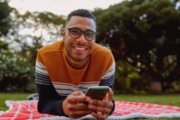 Portrait of a smiling african american young man lying on blanket in the park holding mobile phone in hand looking at camera smiling - Fotografie, Obrázek