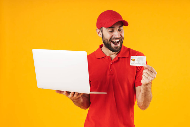 Portrait of handsome delivery man in red t-shirt and cap holding silver laptop computer and credit card - Фото, зображення