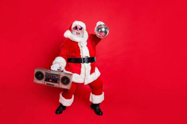 Full length photo of cheerful santa claus in headwear hat holding boom box glittering ball dancing on newyear party wearing style stylish eyeglasses eyewear isolated over red background - 写真・画像