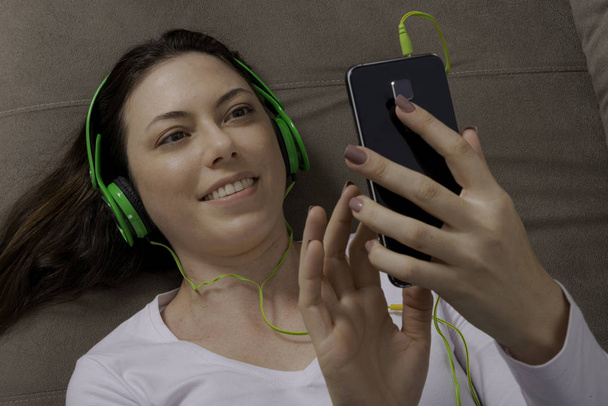 Hermosa mujer relajándose en su sofá escuchando música con su teléfono celular
. - Foto, Imagen