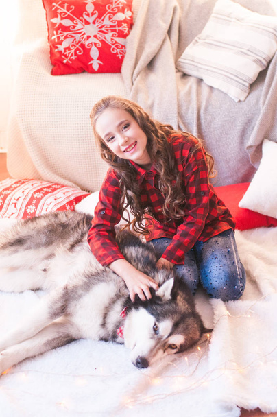 Cute girl in a red checkered shirt with malamute at home in the room decorated for Christmas. Happy christmas mood - Foto, Imagen