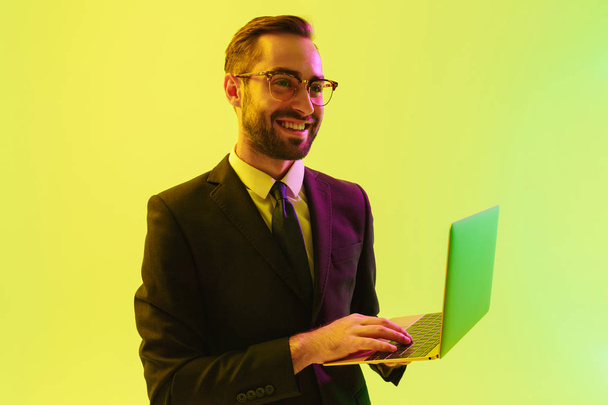 Optimistic pleased positive happy young business man posing isolated over light green background wall with led neon lights using laptop computer. - Valokuva, kuva
