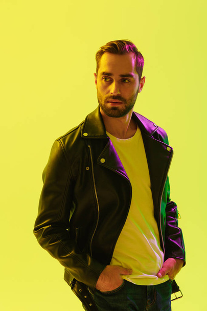 Handsome young stylish man posing isolated over light green background wall with led neon lights dressed in leather jacket. - Foto, imagen