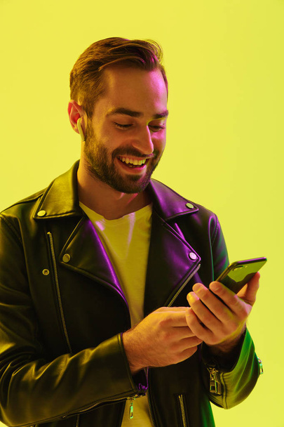 Happy cheerful handsome young stylish man isolated over light green background wall with led neon lights dressed in leather jacket using mobile phone. - Fotoğraf, Görsel
