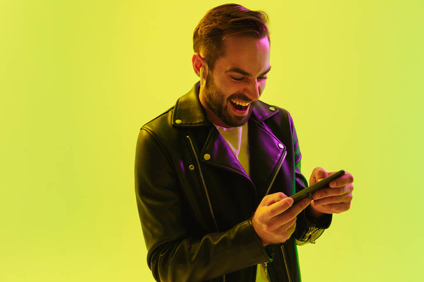 Emotional young stylish man isolated over light green background wall with led neon lights dressed in leather jacket play games by mobile phone. - Foto, afbeelding