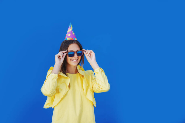 Attractive Caucasian teenage girl in yellow blouse and with birthday cap on head putting on eyeglasses while standing in front of blue background. - Photo, Image