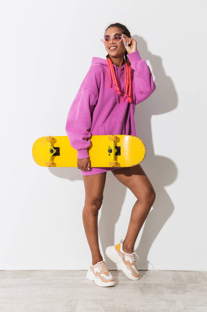 Smiling pleased young african woman posing isolated over white wall background in bright pink sweatshirt holding skateboard. - Фото, изображение