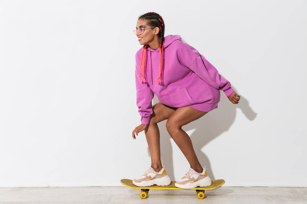 Young african woman skater isolated over white wall background in bright pink sweatshirt on skateboard. - Фото, изображение