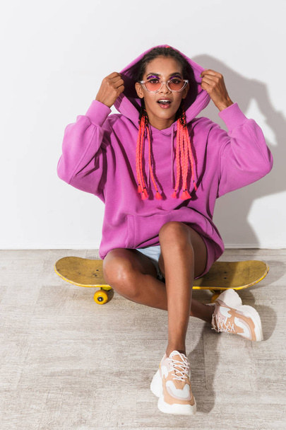 Amazing young african woman isolated over white wall background in bright pink sweatshirt sit on skateboard. - Фото, зображення