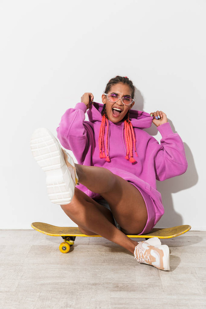 Cheerful smiling optimistic young african woman isolated over white wall background in bright pink sweatshirt sit on skateboard. - Foto, imagen