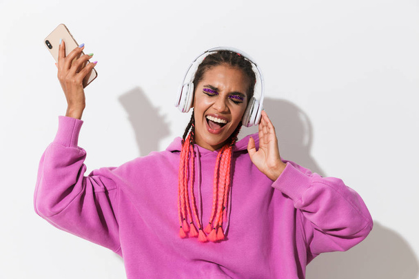 Pleased positive young african woman isolated over white wall background in bright pink sweatshirt using mobile phone listening music with headphones. - Foto, imagen