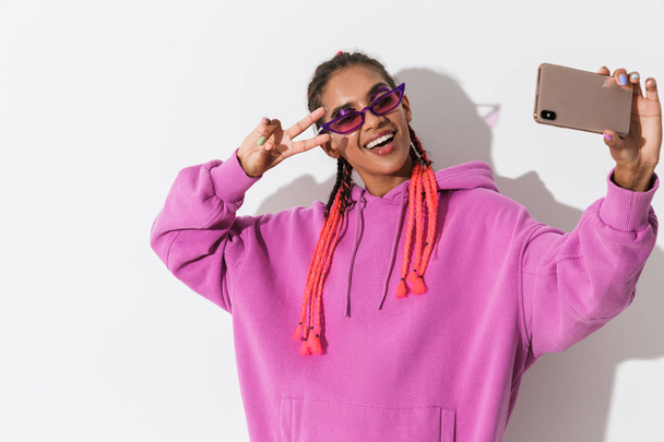 Smiling young african woman isolated over white wall background in bright pink sweatshirt wearing sunglasses take a selfie by mobile phone. - Photo, image
