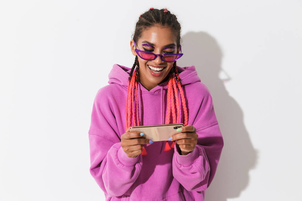 Optimistic happy young african woman isolated over white wall background in bright pink sweatshirt wearing sunglasses play games by mobile phone. - Foto, Imagen