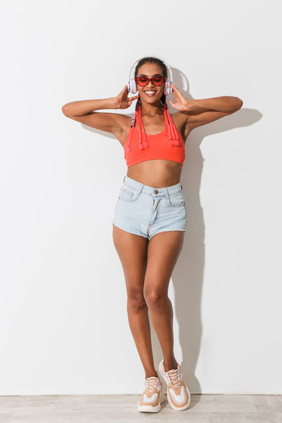 Positive young african stylish woman in sunglasses posing isolated over white wall background listening music with headphones. - Photo, Image