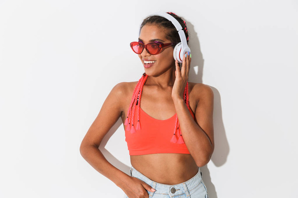 Positive optimistic young african stylish woman in sunglasses posing isolated over white wall background listening music with headphones. - 写真・画像
