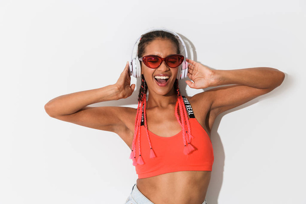 Positive optimistic young african stylish woman in sunglasses posing isolated over white wall background listening music with headphones. - Fotografie, Obrázek
