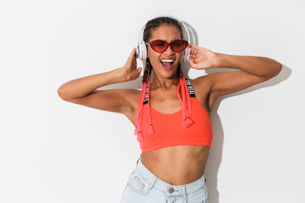 Positive optimistic young african stylish woman in sunglasses posing isolated over white wall background listening music with headphones. - Foto, Imagem