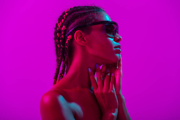 Attractive beautiful young african naked woman posing isolated over purple neon lights wall background wearing sunglasses. - Photo, Image