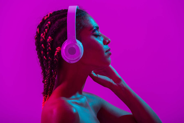 Amazing beautiful young african naked woman posing isolated over purple neon lights wall background listening music with headphones. - Zdjęcie, obraz