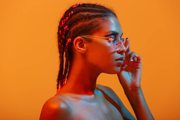 Beautiful young african naked woman posing isolated over yellow neon lights wall background wearing sunglasses. - Φωτογραφία, εικόνα