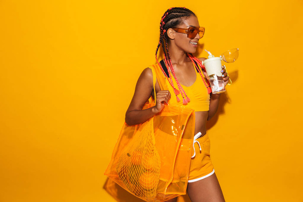 Image of happy african american woman wearing sunglasses carrying bag and holding plastic cup - Photo, image