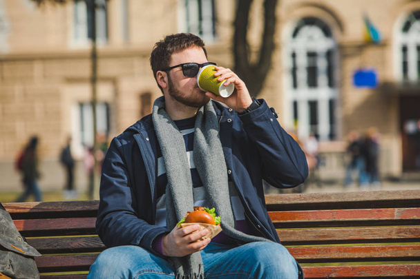 joven sonriente hombre comiendo hamburguesa y beber café sentado en banco de la ciudad
 - Foto, imagen