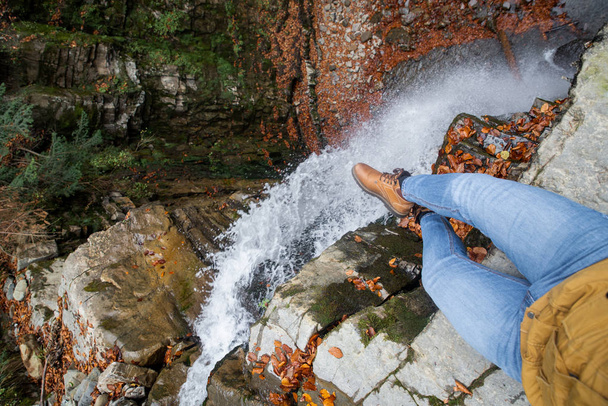 pernas de homem em jeans azuis e botas marrons sentadas na borda olhando para cachoeira
 - Foto, Imagem