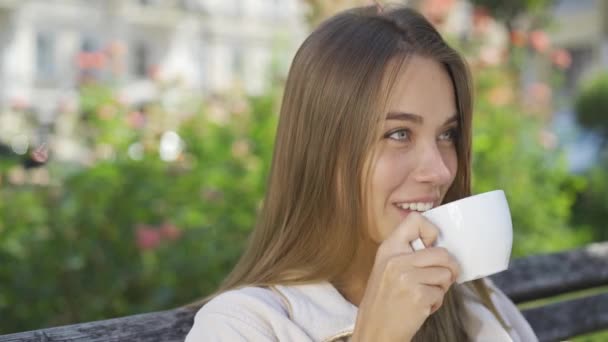 Portrait smiling young woman sitting on the bench in the park drinking coffee or tea. Confident girl enjoying beautiful morning outdoors. Carefree life - Záběry, video