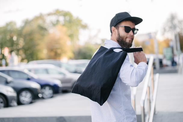 Молодой человек держит черную текстильную эко-сумку на фоне города. - Да. Концепция экологии или охраны окружающей среды. Черная экосумка для насмешек
 - Фото, изображение