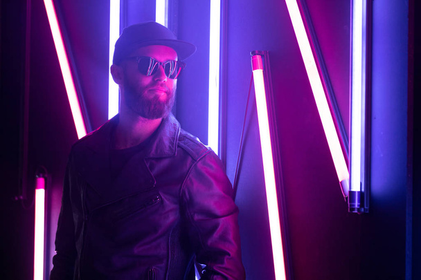 Hipster handsome man on the city streets being illuminated by neon signs. He is wearing leather biker jacket or asymmetric zip jacket with black cap, jeans and sunglasses. - Фото, зображення