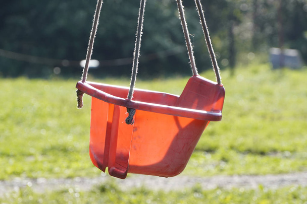 rote Babyschaukel aus Plastik, die an einem sonnigen Tag an einem Baum hängt. Erholung auf dem Land für Kleinkinder und ihre Eltern, Leben im Lieferwagen, Erholung in Zelten und auf Campingplätzen - Foto, Bild