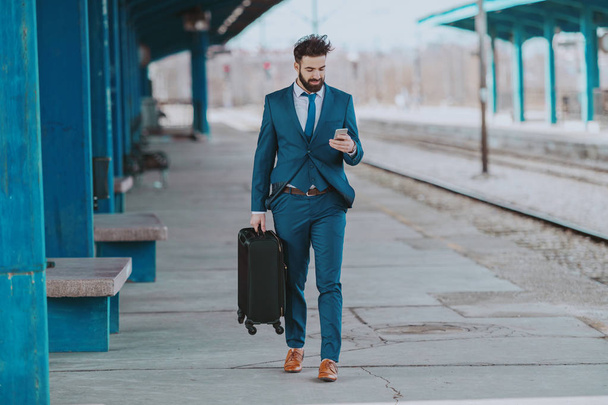Full length of ambitious bearded Caucasian businessman in blue suit carrying luggage and using smart phone while walking at train station. - Zdjęcie, obraz