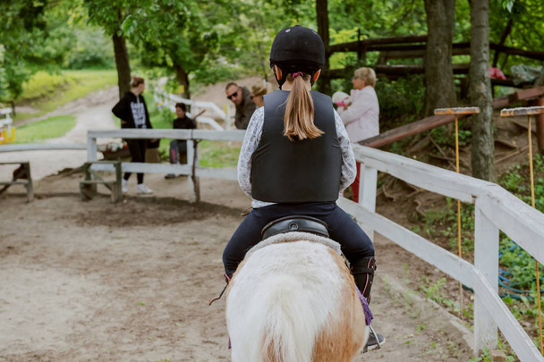 Caucasian girl with helmet and protective vest on riding cute white and brown pony horse. Sunny day on ranch concept. Backs turned. - Photo, Image