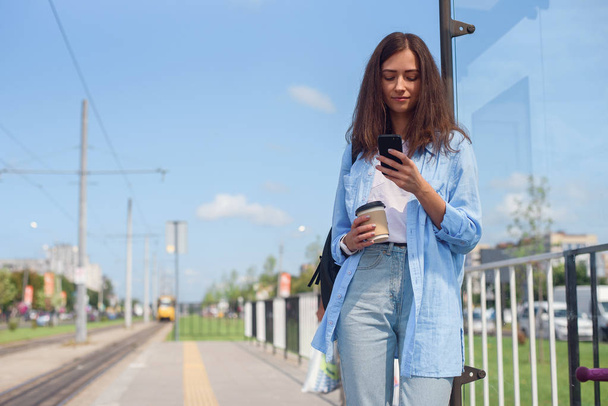 Pretty girl with cup of coffee waits for bus or tram on public transport station in the morning. Young woman with smart phone monitoring transport through the app. - Фото, изображение