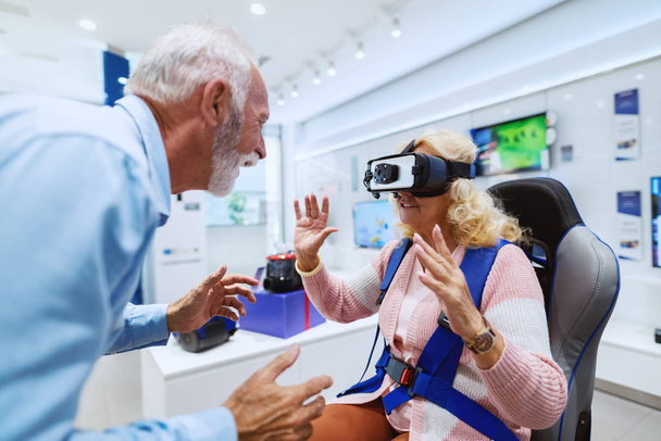 Bonito casal de idosos caucasianos sorrindo experimentando tecnologia de realidade virtual. Mulher sentada na cadeira e usando óculos de proteção. Loja de tecnologia interior
. - Foto, Imagem