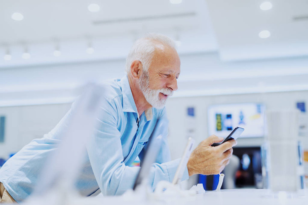 attraktiver lächelnder bärtiger kaukasischer Senior, der sich auf den Stand lehnt und neue Smartphones ausprobiert. Innenausstattung eines Tech-Stores. - Foto, Bild