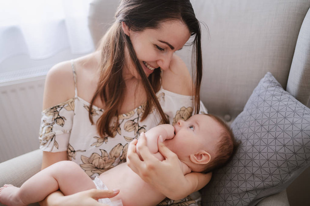 Caring young Caucasian mother holding her 6 months old son holding in arms while sitting in armchair. Baby looking mother in eyes and putting fingers in mouth. - Fotografie, Obrázek