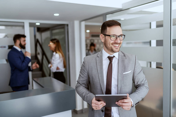 Portrait of handsome Caucasian smiling unshaven businessman in suit and with eyeglasses standing in hall with tablet in hands. In background his colleagues chatting. - 写真・画像