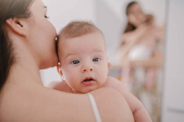 Close up of Caucasian mother holding and kissing her loving six months old son. Baby looking at camera with confused facial expression. - Zdjęcie, obraz