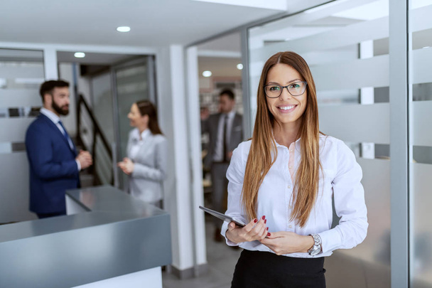 Charming Caucasian businesswoman dressed in shirt and skirt standing on hallway and holding tablet. In background her colleagues standing and chatting. - Foto, afbeelding