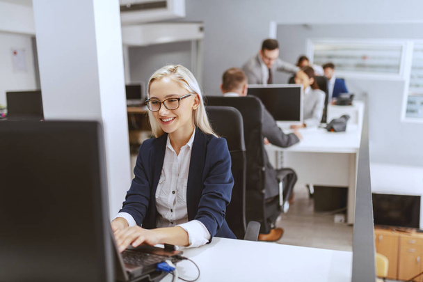 笑顔のブロンド白人ビジネスウーマンは、彼女の職場に座って、コンピュータを使用しています。キーボードでのハンズオン. - 写真・画像