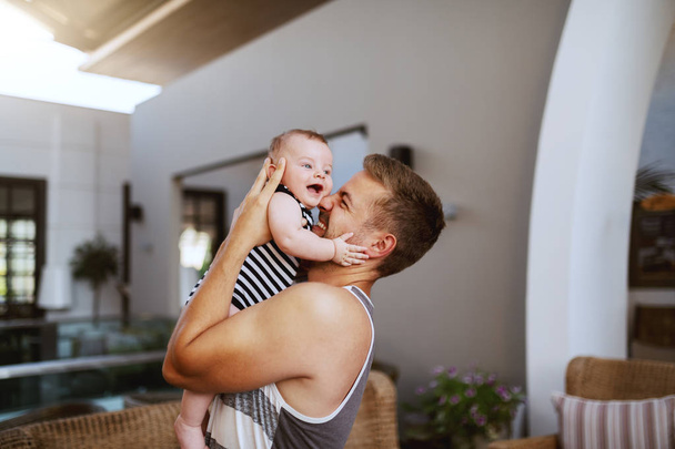 Gelukkig knappe Kaukasische vader knuffelen zijn liefdevolle 6 maanden oude zoon terwijl staande binnen. Baby is lachen en knuffelen vader. - Foto, afbeelding