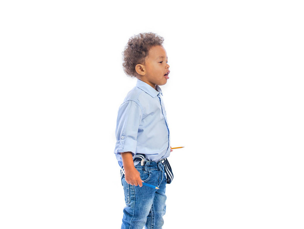 Boy with curly hair in jeans and a light-colored shirt is standing in a half-turn holding a pencils in both hands and looking to the side - Φωτογραφία, εικόνα